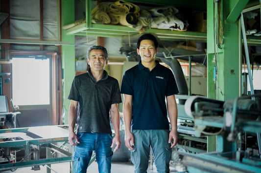 Father and son, Ryushi, smiling as they stand side by side in a tatami workshop with tools and materials in the background.