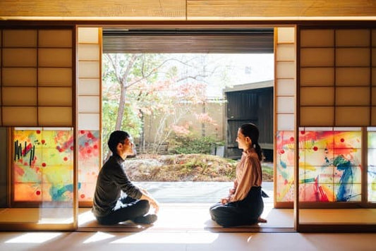 Two people sitting on a tatami mat in a traditional Japanese room, with sliding doors open to a peaceful garden view.